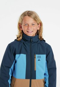 Blå och brun skidjacka med en dragkedjeförsedd bröstficka. Har en huva, varumärkeslogotyp och en färgblockeringsdesign med olika texturer.