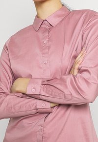 Light pink button-up shirt with a collar, long sleeves, and matching buttons. Smooth fabric with a subtle sheen and a single chest pocket.