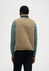 Μπεζ γιλέκο από φλις με ψηλό γιακά, υφασμάτινη υφή και αντίθετο σκούρο περιθώριο. Φοριέται πάνω από ένα πράσινο μακρύ μανίκι μπλουζάκι. Ορατά μαύρα παντελόνια.