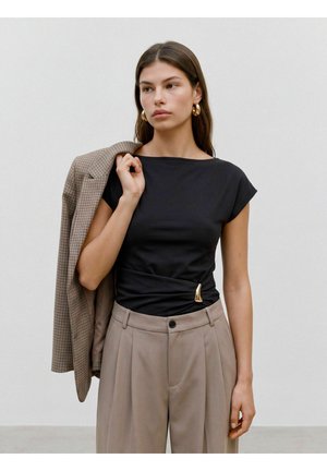 Femme aux longs cheveux bruns portant des boucles d'oreilles créoles dorées, un haut noir, un pantalon beige plissé, et tenant une veste à carreaux marron sur son épaule.