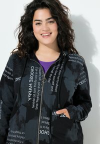 Sudadera con cremallera negra, con texto impreso y patrones florales. Cuenta con capucha ajustable y bolsillos. La tela tiene una textura suave.