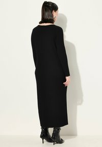 Schwarzes, geripptes Strickkleid mit langen Ärmeln und gerader Silhouette, ausgestattet mit einem runden Ausschnitt und seitlichen Schlitzen. Kombiniert mit schwarzen, samtigen Ankle-Boots mit Absatz.