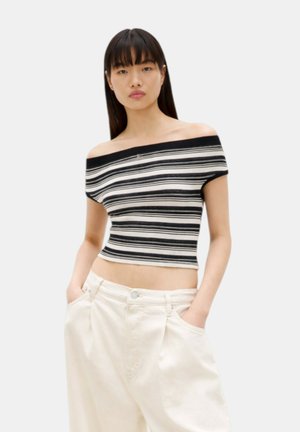 Donna con crop top a righe bianche e nere senza spalle e pantaloni bianchi larghi, mani nelle tasche, in piedi davanti a uno sfondo semplice.