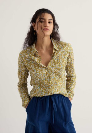 Young woman with long curly hair wearing a yellow floral buttoned shirt and loose navy blue pants, hands in pockets, neutral background.