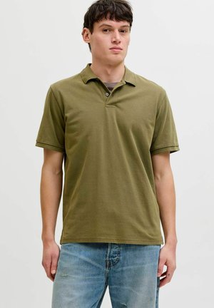 Mann iført olivengrønn kortermet polotrøye over grå undertrøye og lyseblå jeans, stående mot en ensfarget hvit bakgrunn.