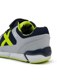 Zapato deportivo infantil con malla gris y azul marino, acentos en amarillo brillante, tira de Velcro y suela texturizada para mayor agarre.