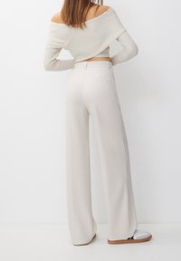 Vrouw met haar rug naar de kijker in een off-shoulder ribgebreide witte trui, hoge taille wijde crèmekleurige broek en witte sneakers met rubberen zolen.
