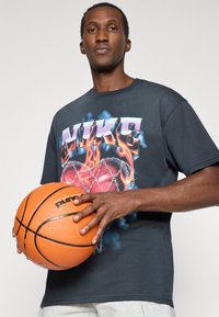 Μαύρη βαμβακερή μπλούζα T-shirt με πολύχρωμο λογότυπο Nike με φλόγες, φορεμένη από ένα άτομο που κρατάει μια πορτοκαλί μπάλα μπάσκετ με υφή.