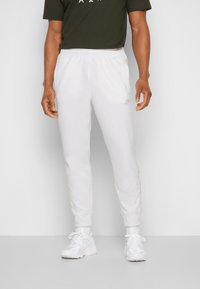 Bílé sportovní tepláky z měkkého materiálu s elastickým pasem a postranními kapsami, v kombinaci s bílými teniskami.