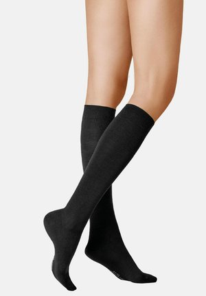 Beine mit schwarzen knielangen Kompressionssocken vor einem einfarbigen weißen Hintergrund, die Passform und Abdeckung der Socken zeigend.