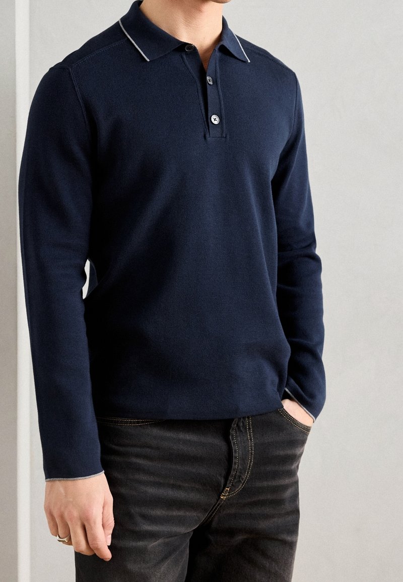Homme portant un polo bleu marine à manches longues avec un liseré blanc, associé à un jean noir, debout avec une main dans la poche.