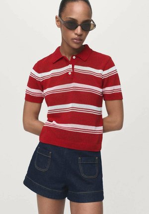 Persoon met donkere zonnebril, rood-wit gestreept kort mouw poloshirt en donkere spijkerbroek met voorzakken, staande met de handen achter de rug.