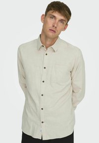 Camisa de manga larga color beige claro, con botones delanteros de color marrón y un bolsillo en el pecho, confeccionada con tecido texturizado.