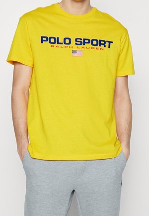 Camiseta amarilla de manga corta con el texto "POLO SPORT RALPH LAUREN" y una pequeña bandera americana, combinada con pantalones deportivos gris claro.