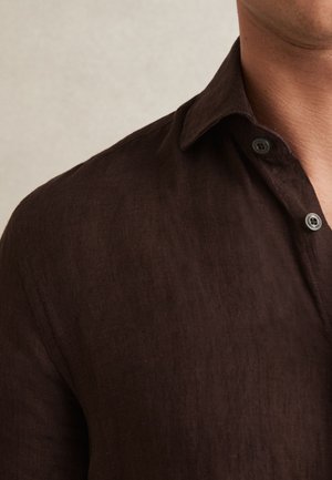 Man draagt een donkerbruin, knoop-overhemd met kraag tegen een effen lichte achtergrond, met zicht op schouder- en nekgebied.
