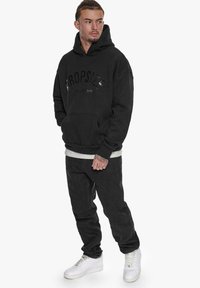 Svart hettegenser med en glanset "DROP SIZE LIMITED EDITION" logo, kombinert med mørke bukser og hvite sneakers. Har en frontlomme.
