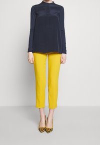 Donkerblauwe blouse met lange mouwen en hoge kraag, gecombineerd met felgele op maat gemaakte broek en geprinte gele en zwarte hoge hakken.