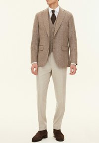 Brauner Wollmischblazer mit Reverskragen, Drei-Knopf-Verschluss und zwei vorderen Taschen, kombiniert mit einer hellbeigen Weste und Hose. Braune Wildlederschuhe.