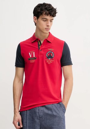 Polo rosso con maniche blu scuro, loghi ricamati e dettagli in patch. Maniche corte, colletto e design a orlo dritto.