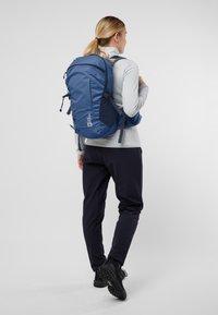 Blauer Rucksack mit gepolsterten Trägern, verstellbarem Design und einem auffälligen Logo. Glattes Material mit einer eleganten, schlanken Form.