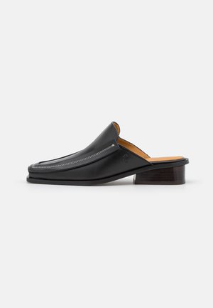 Mules de cuero negro con diseño curvado, que presentan costuras blancas y un tacón de block de madera. Textura suave con un estilo minimalista.