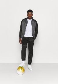 Sportset bestaande uit een grijs en zwart jack, een wit shirt en zwarte broek. Opvallende gouden voetbal en witte sneakers met groene accenten.