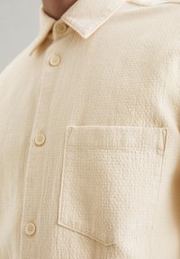 Beige button-up shirt with a textured fabric, featuring a collar and a single chest pocket. Buttons are beige with a smooth finish.