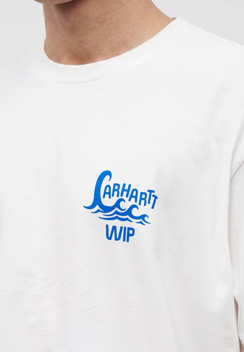 Maglietta bianca con scritta blu "Carhartt WIP" e motivo a onda sul petto sinistro, indossata da una persona mostrata dal collo alla parte superiore del busto.
