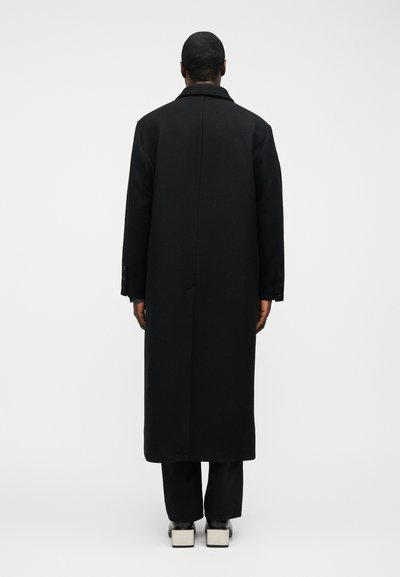 Άτομο που στέκεται με την πλάτη, φοράει μακρύ μαύρο παλτό, μαύρο παντελόνι και παπούτσια με τετραγωνισμένη φτέρνα σε άσπρο και μαύρο, σε απλό φόντο.