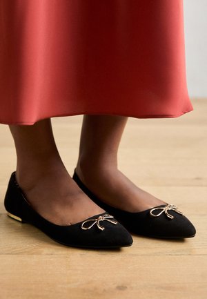 Czarne zamszowe płaskie buty z wąskim noskiem, z metalowym akcentem w postaci kokardy na przodzie i złotym detalem na pięcie.