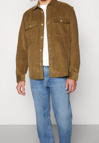Chaqueta de pana marrón con dos bolsillos en el pecho, cierre de botones en la parte delantera y un acabado de textura suave, combinada con unos jeans de color azul claro y una camisa blanca.