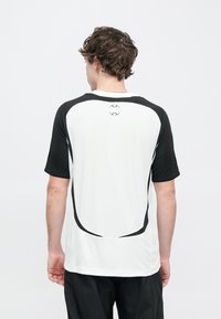 Koszulka sportowa z krótkim rękawem, biała na tułowiu, z czarnymi rękawami i zakrzywionym czarnym szwem ozdobnym. Na karku znajduje się okrągłe logo.