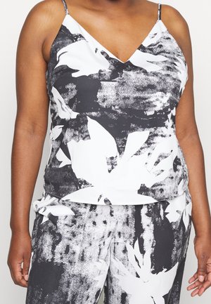 Femme portant un haut caraco sans manches à motif floral noir et blanc avec un pantalon assorti sur un fond uni.