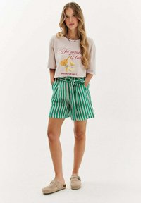 Krémové grafické tričko s květinovým vzorem a červeným textem, v kombinaci se zelenými a bílými proužkovanými kraťasy staženými v pase a béžovými klouzavými botami.