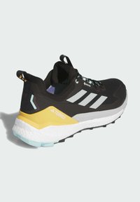 Chaussure de trail running noire avec des accents jaunes et teal, présentant une tige texturée, une semelle rembourrée et un logo à trois bandes bien marqué.