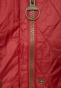 Czerwona pikowana kurtka z centralnym zamkiem błyskawicznym z okrągłym metalowym uchwytem z tłoczonym napisem "Barbour" oraz zatrzaskami u dołu.
