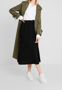 Cappotto trench verde oliva sopra una camicia bianca con bottoni, abbinato a una gonna nera lunga fino alla caviglia e sneakers bianche. Texture morbide, vestibilità classica.
