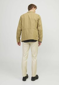 Leichte, khakifarbene Jacke mit lockerer Passform, Taillierung, langen Ärmeln und geradem Saum. Getragen mit cremefarbenen Hosen und schwarzen Schuhen.
