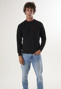 Mustan kapuutsiga sviiter, millel on pikad raglan-sleeved varrukad, soonikust pöidlad ja serv, millel on väike punane logo. Koos helesiniste denim-teksadega.