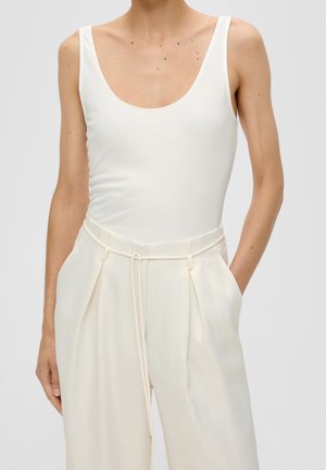 Person trägt ein ärmelloses weißes Tanktop, das in eine hoch taillierte, cremefarbene Hose mit dünnem Gürtel gesteckt ist, die Hände in den Taschen.