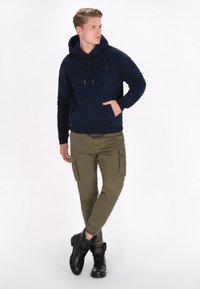 Námořnická mikina s kapucí, kapsou a šňůrkami, doplněná olivovými cargo kalhotami s bočními kapsami a černými kotníkovými botami.