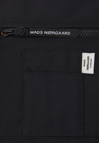 Black fabric bag with a zippered pocket. Features a rectangular patch and a brand label in white, highlighting stitching and material texture.