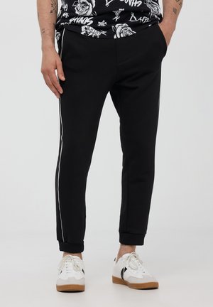 Pantaloni sportivi neri con un design affusolato, strisce laterali bianche e polsini a coste. Indossati con delle scarpe da ginnastica bianche.