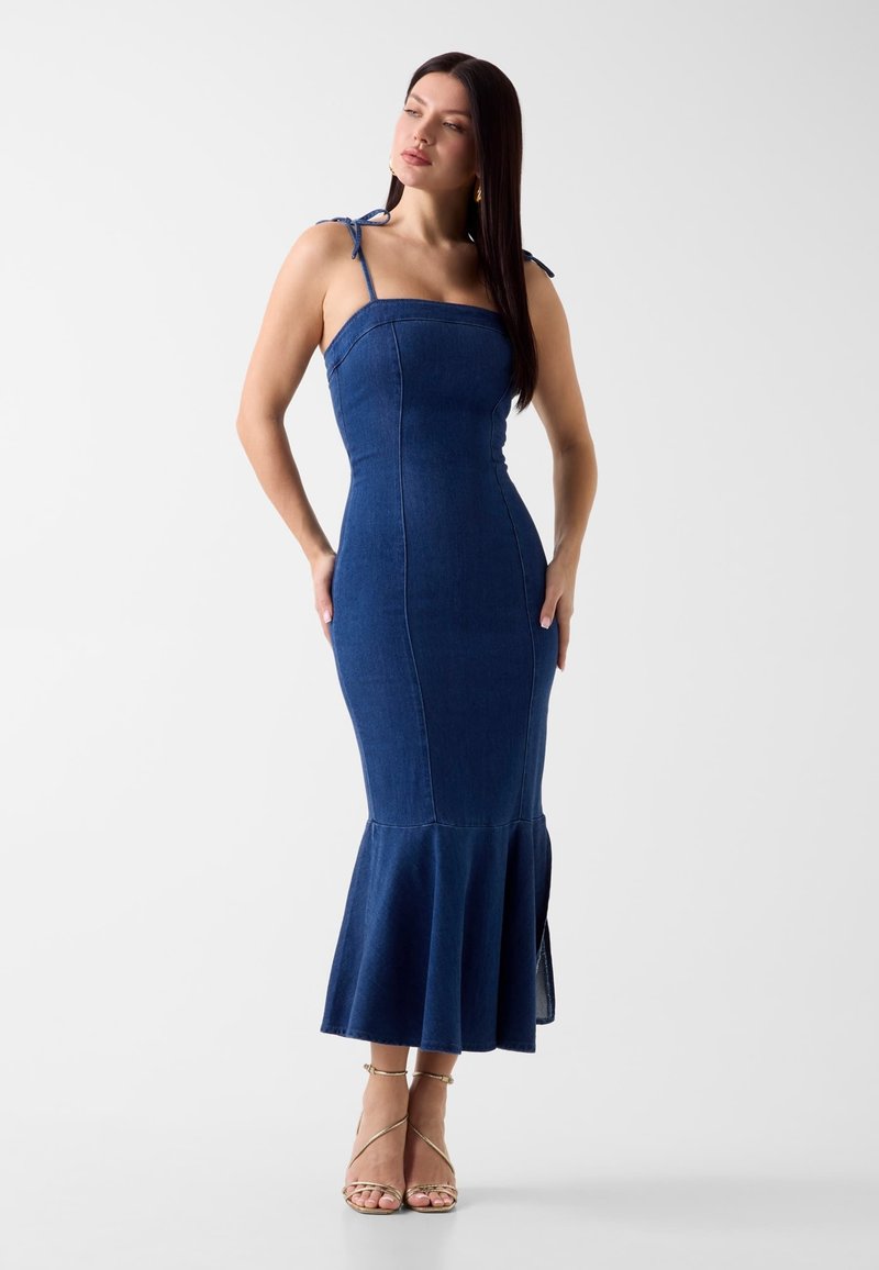 Femme portant une robe en denim bleu foncé moulante sans manches avec des bretelles à nouer et un ourlet évasé, associée à des talons dorés à lanières, debout sur fond blanc.