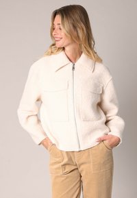 Witte, fluffy zip-up jas met twee borstzakken, kraagontwerp en textuurstof, gecombineerd met beige corduroy broek.