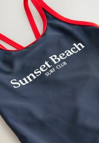 Granatowy strój kąpielowy z czerwonymi ramiączkami, na którym znajduje się napis "Sunset Beach SURF CLUB" w białym kolorze. Gładki, elastyczny materiał.