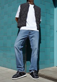 Chaleco negro acolchado, camiseta blanca, jeans azules y zapatos negros sin cordones con suelas blancas, contra un fondo de pared de azulejos color teal.