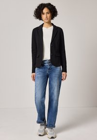 Femme aux cheveux bouclés portant un blazer noir texturé, un T-shirt blanc, un jean bleu et des baskets grises, debout devant un fond uni.