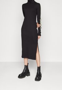 Svart långärmad klänning med polokrage, sidoklyftor och ett grönt rand längs ärmarna, parat med kraftiga svarta ankelboots.