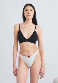 Czarny bralet z cienkimi ramiączkami i dekoltem w kształcie litery V, połączony z białymi stringami z czerwonym akcentem na metce. Gładka tkanina, minimalistyczny styl.